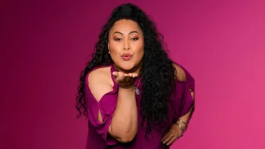 Woman with long curly hair blows a kiss against a pink background.