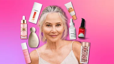 A smiling older woman surrounded by various skincare products on a pink gradient background.