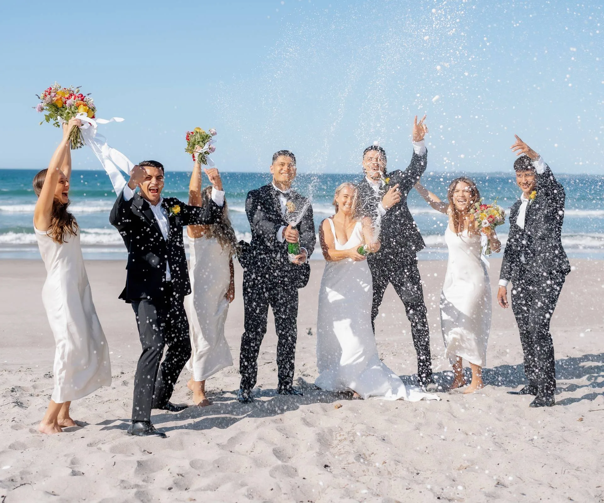 Popping champagne with their bridesmaids and groomsmen on a beach