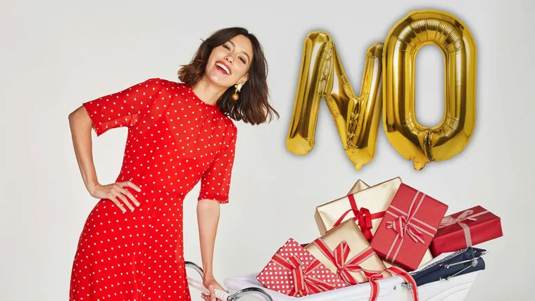 Woman in red dress holding a trolley with presents. Big gold helium balloons behind her spelling out 'NO'