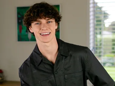 Austin Taylor smiling from a stool in his living room