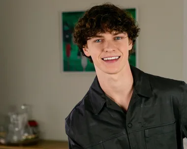 Austin Taylor smiling from a stool in his living room