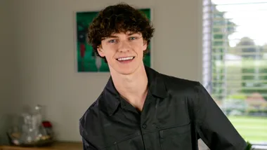 Austin Taylor smiling from a stool in his living room