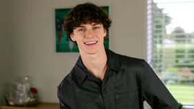 Austin Taylor smiling from a stool in his living room