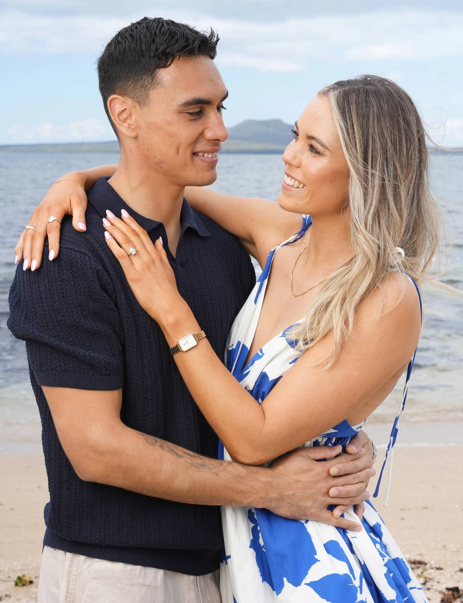 The couple smiling in front of a view across the water of Rangitoto Island, with Greer's hand on Max's chest, showing off the ring