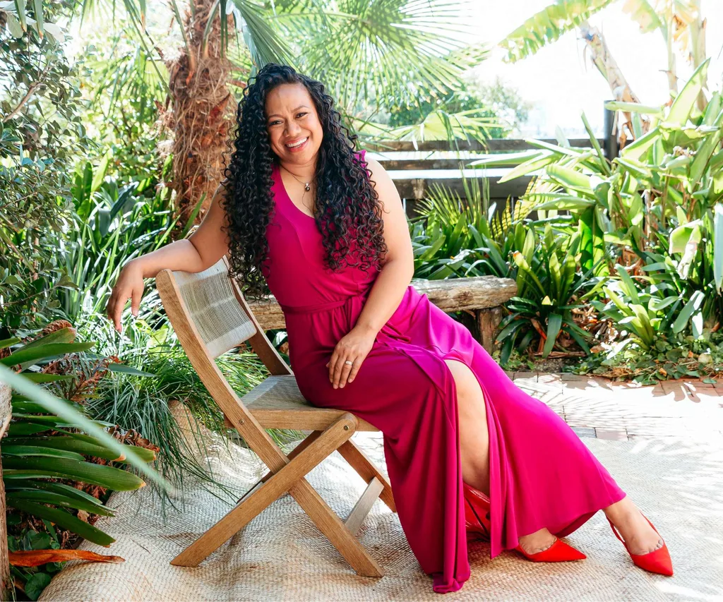 Indira Stewart wearing a red dress leaning on a chair in front of some lush greenery.