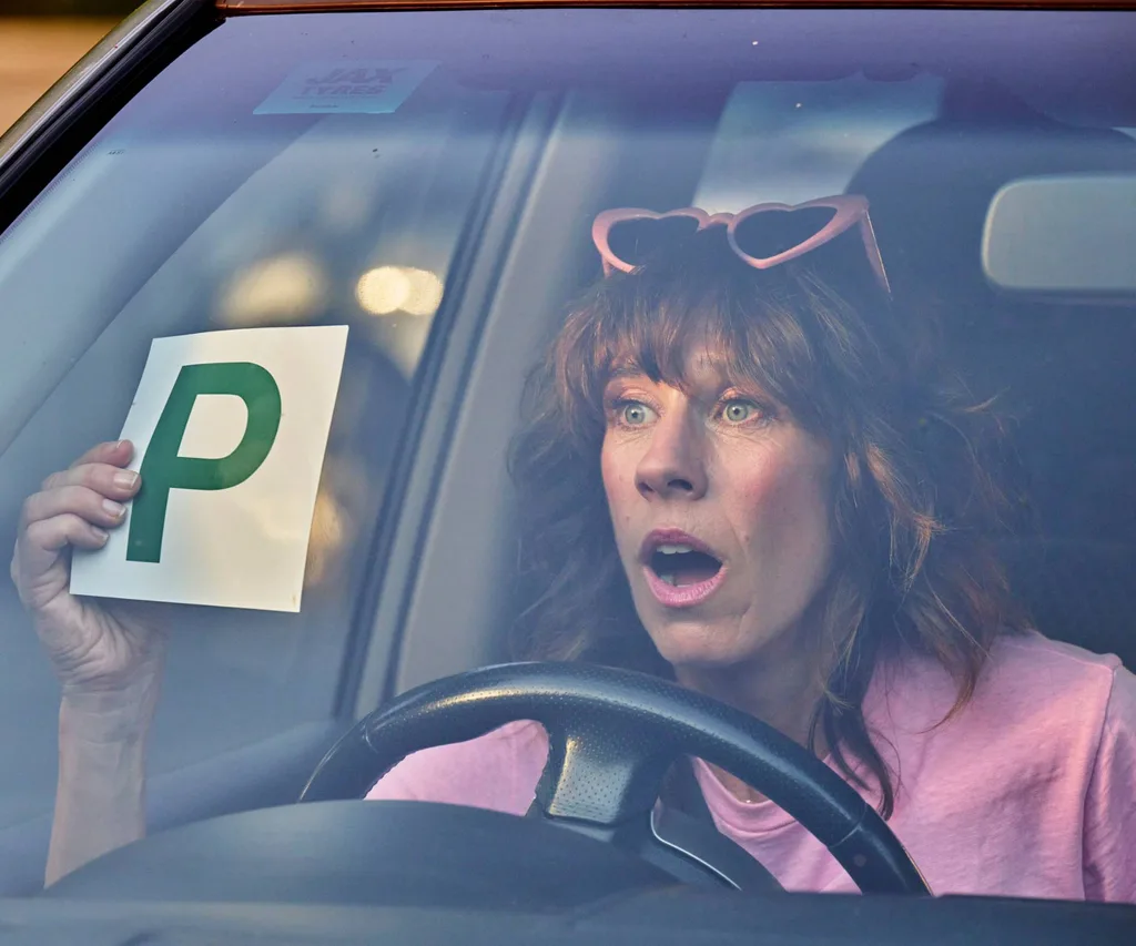 woman in car wearing a pink jacket and pink glasses holding a P plate