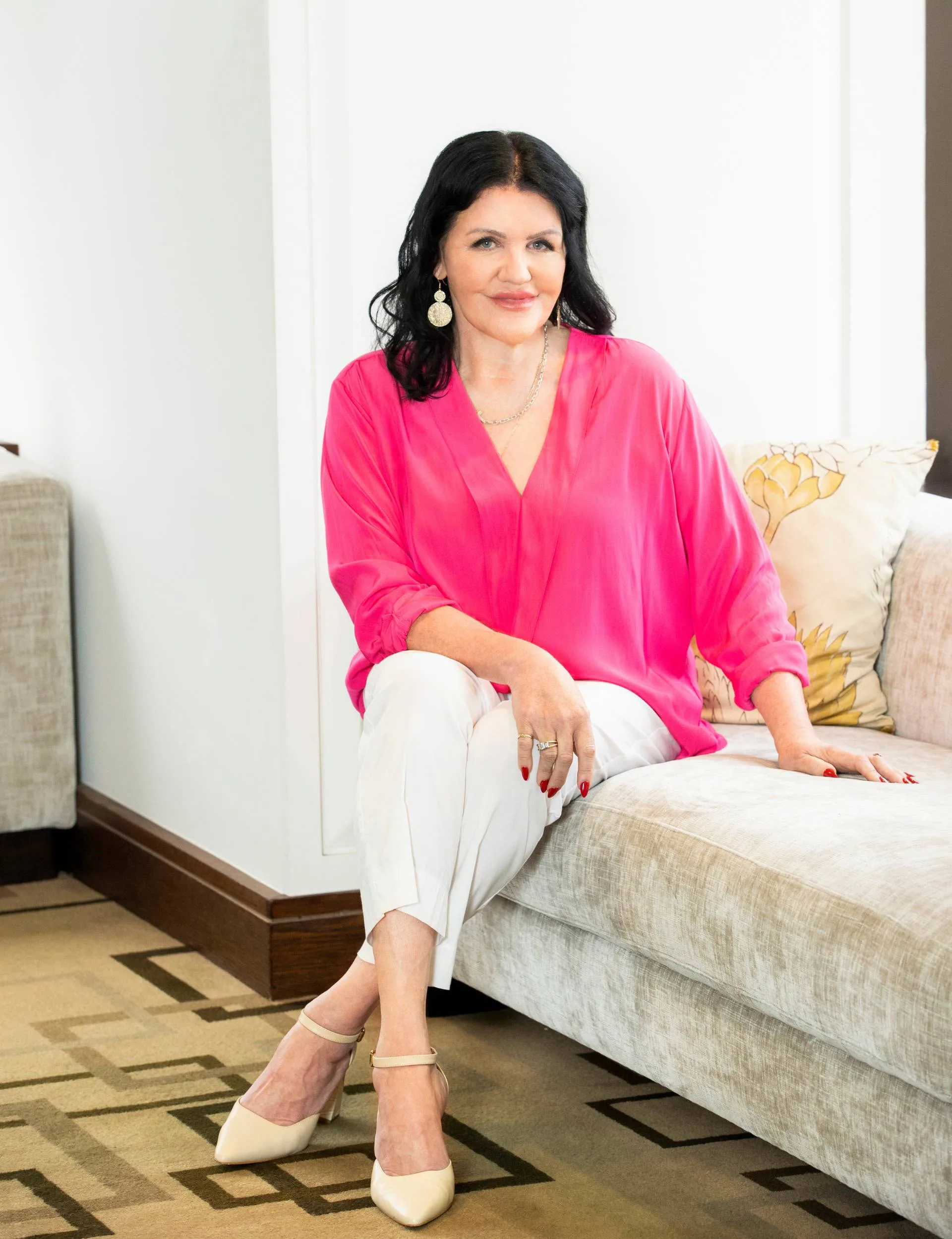 Polly Gillespie sitting on a couch in a fluro pink shirt and white pants