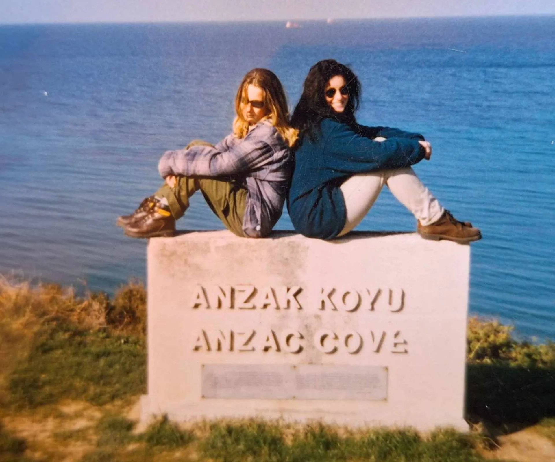 Alexandra Lattey sitting on an 'ANZAC COVE' plaque with a friend