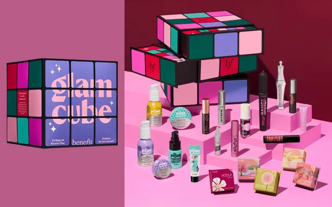 A colourful display of beauty products is arranged with a Rubik's Cube-themed box labeled "glam cube." The products include mini bottles and boxes, set against a pink and red background.