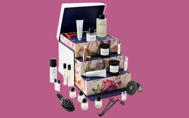 A luxurious beauty advent calendar with patterned drawers displaying various skincare and fragrance products, brushes, and tools on a purple background.