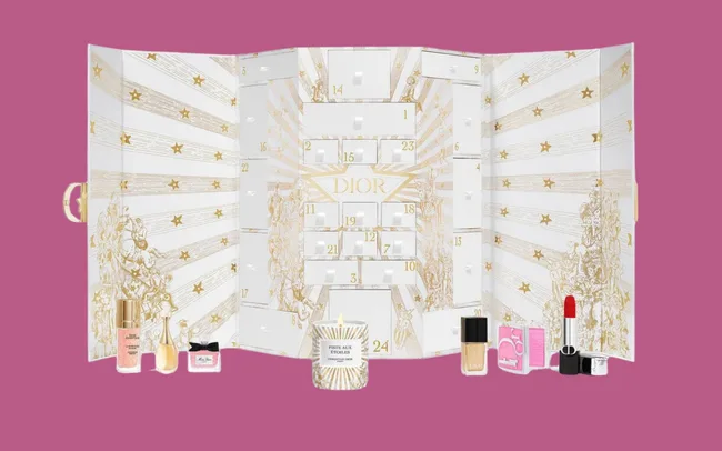 Luxurious Dior advent calendar with 24 numbered slots on gold starry panels. Displayed in front are various cosmetics, including lipstick and perfume, on a pink background.
