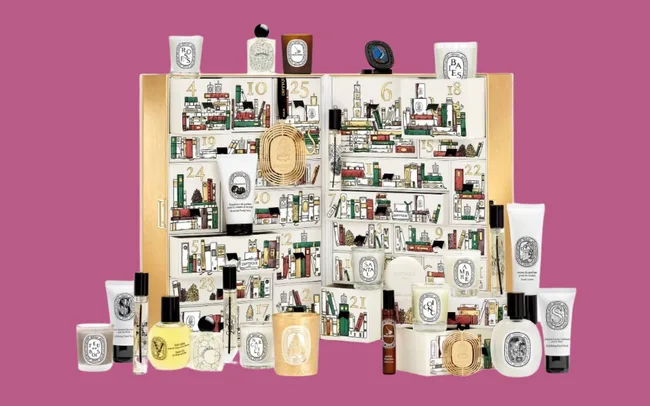 Diptyque Advent calendar with illustrated cityscape design, surrounded by assorted scented candles, perfumes, and skincare products on a pink background.
