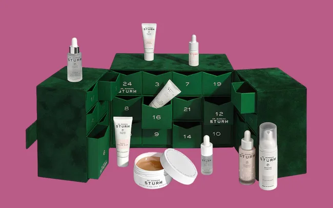 Green skincare advent calendar with products on a pink background. Boxes numbered 1-24; various serums, creams, and bottles are displayed. Luxurious and festive.