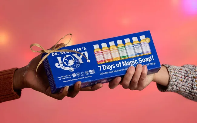 Two hands exchange a blue box labeled "7 Days of Magic Soap" by Dr. Bronner's against a soft pink backdrop. The mood is celebratory and gifting.