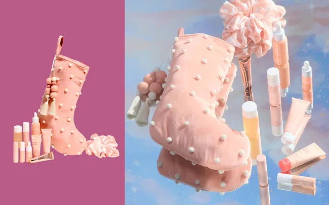 A pink stocking adorned with white pom-poms is surrounded by various skincare products and a scrunchie, set against a soft, pastel background.