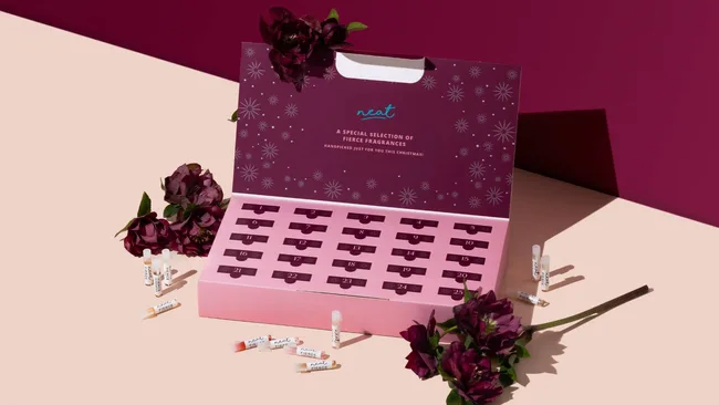 Elegant fragrance advent calendar with burgundy and pink design, labeled "A special selection of fine fragrances." Surrounded by dark flowers and small perfume vials.