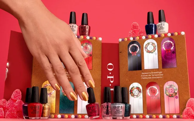 A hand with gold nails reaches for a festive OPI nail polish advent calendar set against a red backdrop, surrounded by vibrant nail polish bottles.