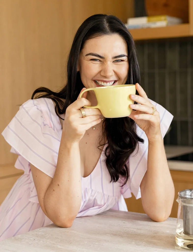 Woman in kitchen in pink shirt, drinking out of a yellow mug