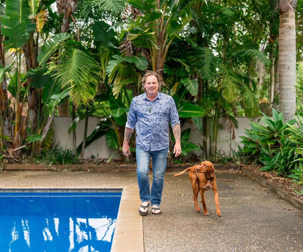 Man in blue shirt, jeans and sandals walking with brown dog by the pool