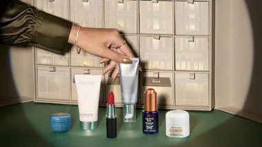A woman's hand reaches for a silver tube of serum among luxury cosmetics on a green surface. Background features a decorative advent calendar. Elegant and sophisticated.
