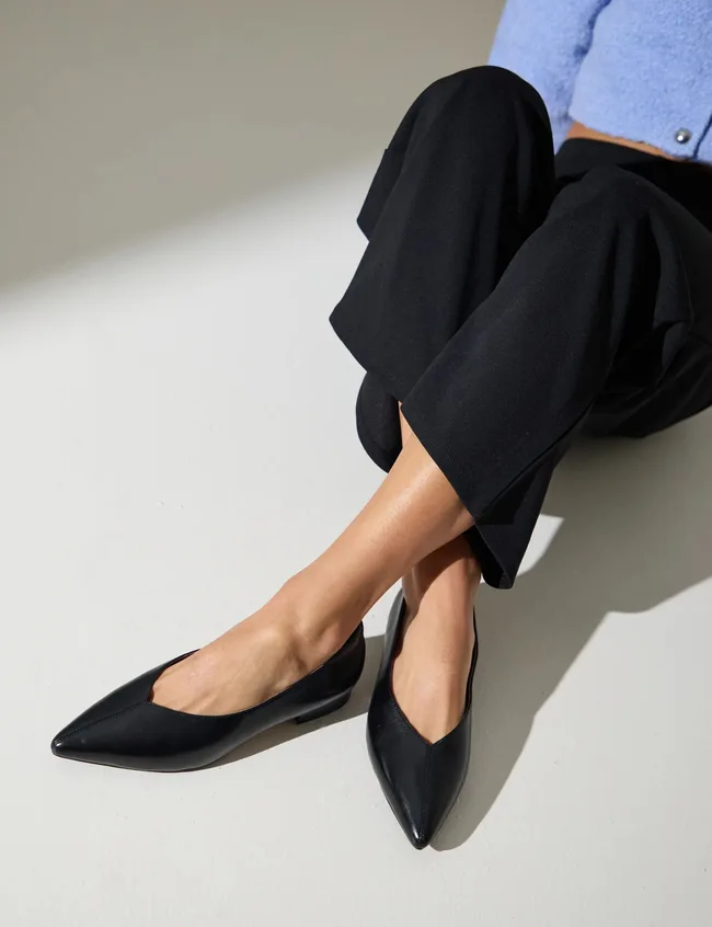 Woman sitting on floor, cropped from waist down, shown wearing black trousers and black pointed toe flat shoes