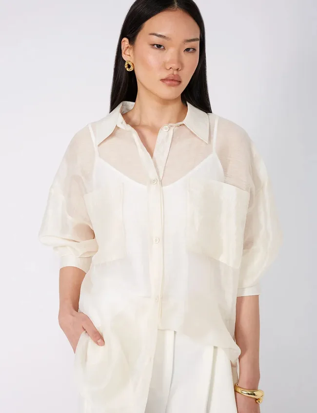 Crop of woman from waist up, wearing a sheer, short-sleeved cream button-up shirt over a white camisole