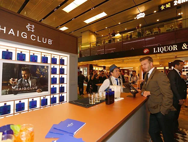"Bar counter at Haig Club, with a large display, bottles and two men conversing, one in a suit, indoor setting with onlookers."