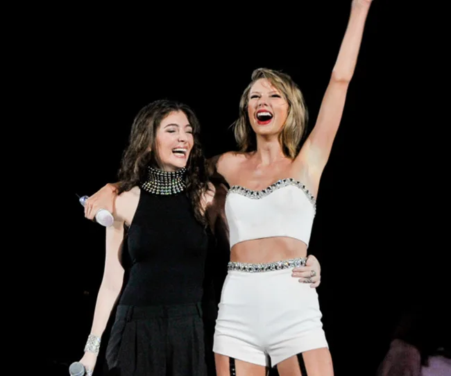 Two women embrace on stage, smiling, one raising an arm. They wear fashionable outfits and hold microphones.
