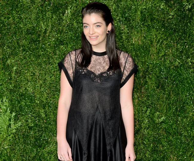 A person with long dark hair in a black lace dress stands in front of a leafy green wall, smiling.