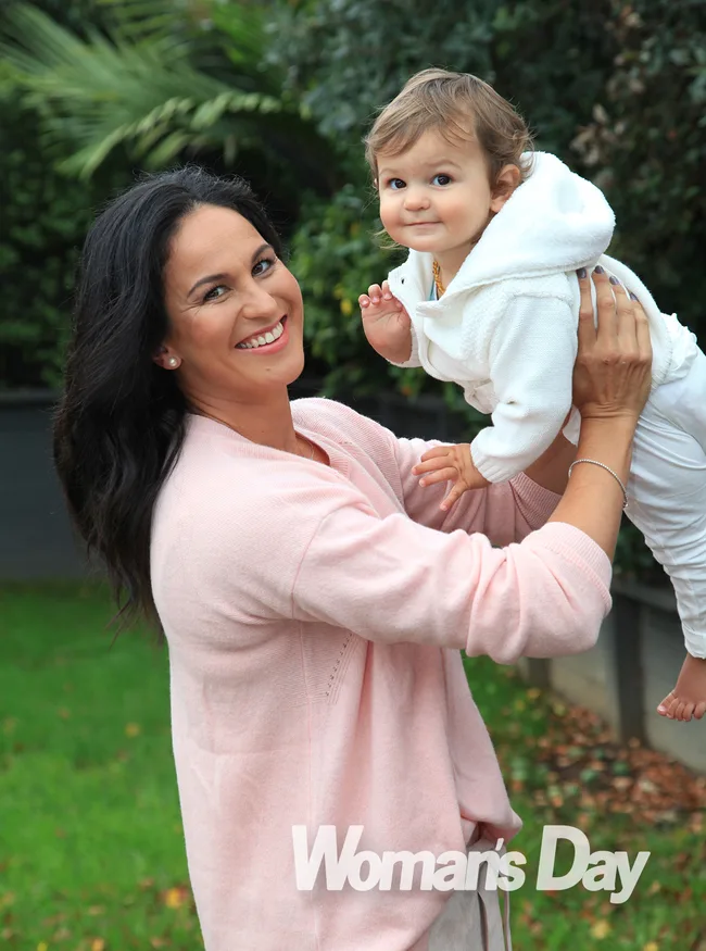 Woman in a pink sweater holding a smiling baby in a white outfit outside, with "Woman's Day" text at the bottom.