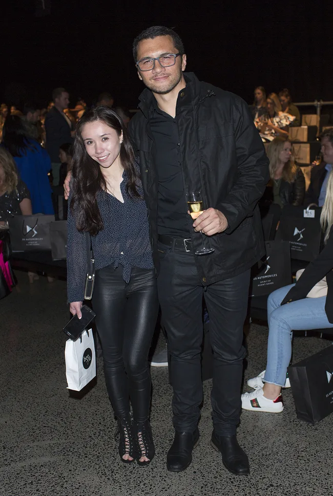 Two people posing together at an event; one holding a champagne glass, the other a shopping bag and phone.