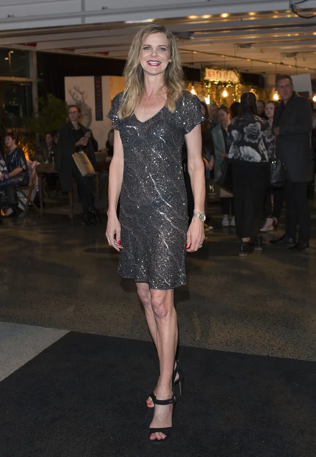 A woman in a sparkly silver dress and black heels poses at a formal event with people in the background.