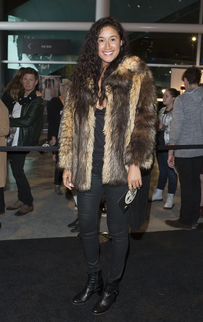 Woman in a fur coat and black pants at an event, smiling at the camera in a busy indoor setting.