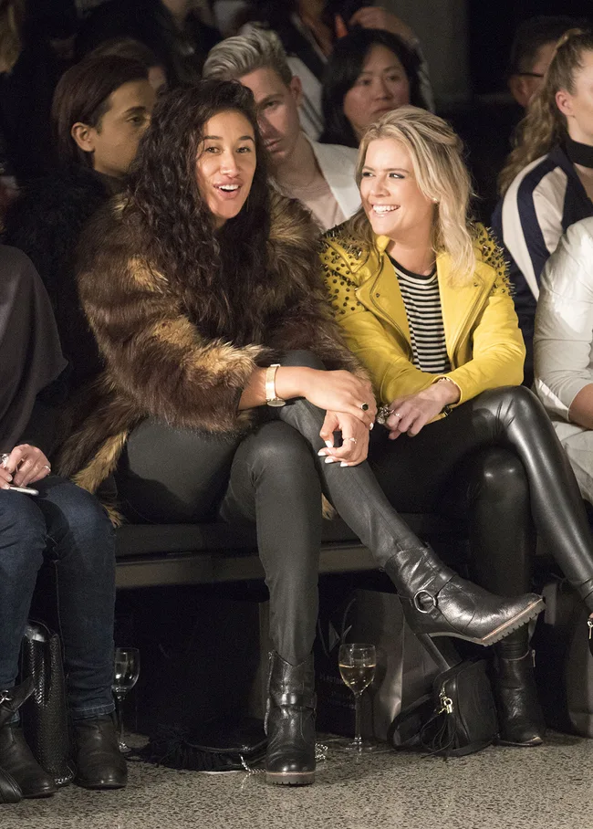 Two women sitting in the front row of a fashion show, one in a fur coat and the other in a yellow studded jacket.