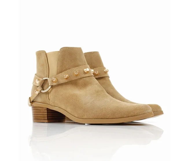 Tan suede ankle boots with studded straps and low wooden heels, displayed on a reflective white surface.
