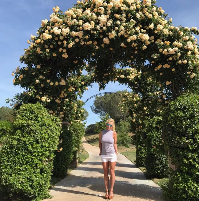 Woman in white outfit standing under an archway of blooming yellow roses on a sunny day.