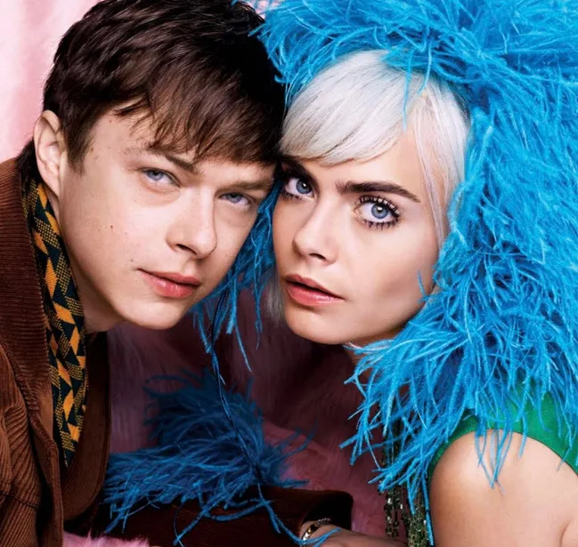 Two people pose closely together, one wearing a blue feathered outfit against a soft pink background.