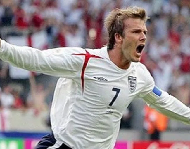 A soccer player in a white England jersey with number 7 celebrates with arms outstretched on the field.