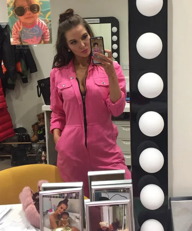 A woman in a pink jumpsuit takes a mirror selfie in a brightly lit room, with framed photos and a child's portrait visible.