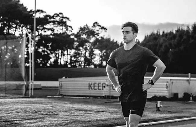 Man standing with hands on hips on a sports field, trees in background, wearing a T-shirt and shorts, black and white image.