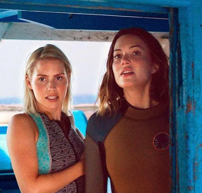 Two women in wetsuits on a boat, looking ahead with expressions of focus, blue ocean visible in the background.
