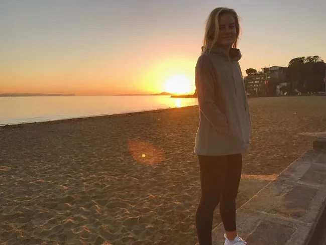 Person standing on a beach during sunset, wearing a gray jacket, with the sun setting over the ocean in the background.