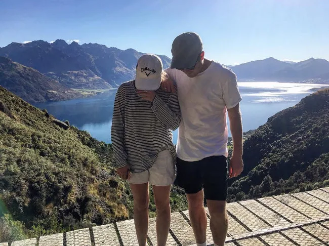A couple stands on a scenic mountain overlook beside a lake, wearing casual clothes and hats on a sunny day.