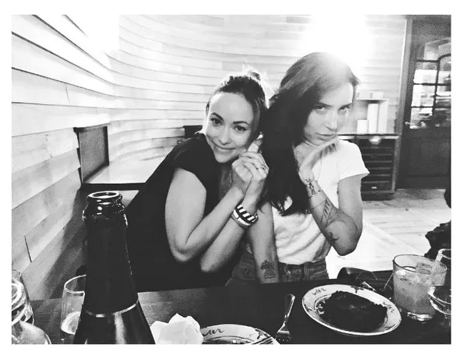 Two women sitting at a dining table, one smiling and the other playfully posing, with food and drinks visible.