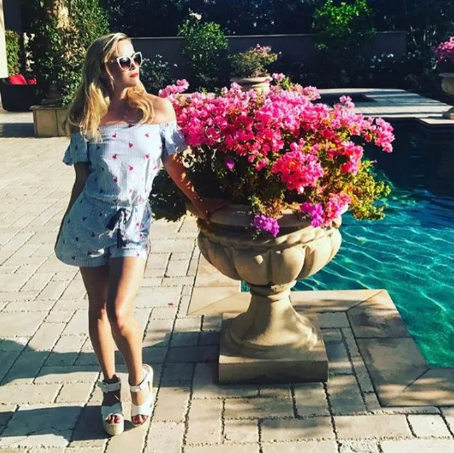 Woman in a floral romper and sunglasses stands by pink flowers near a pool on a sunny day.