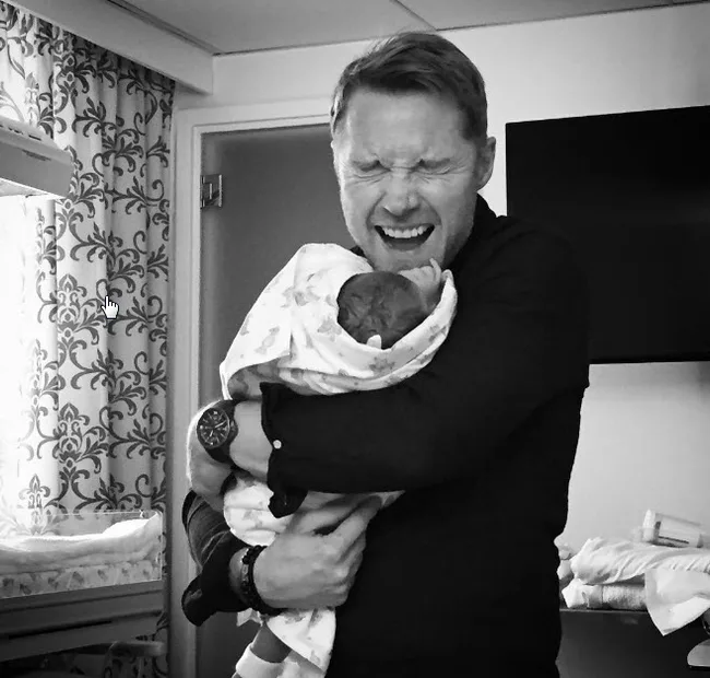 Man joyfully holding a newborn wrapped in a blanket, eyes closed, standing in a room with patterned curtains.