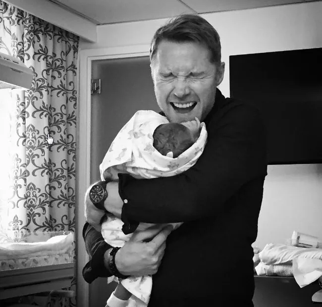 Man joyfully holding a newborn wrapped in a blanket, eyes closed, standing in a room with patterned curtains.