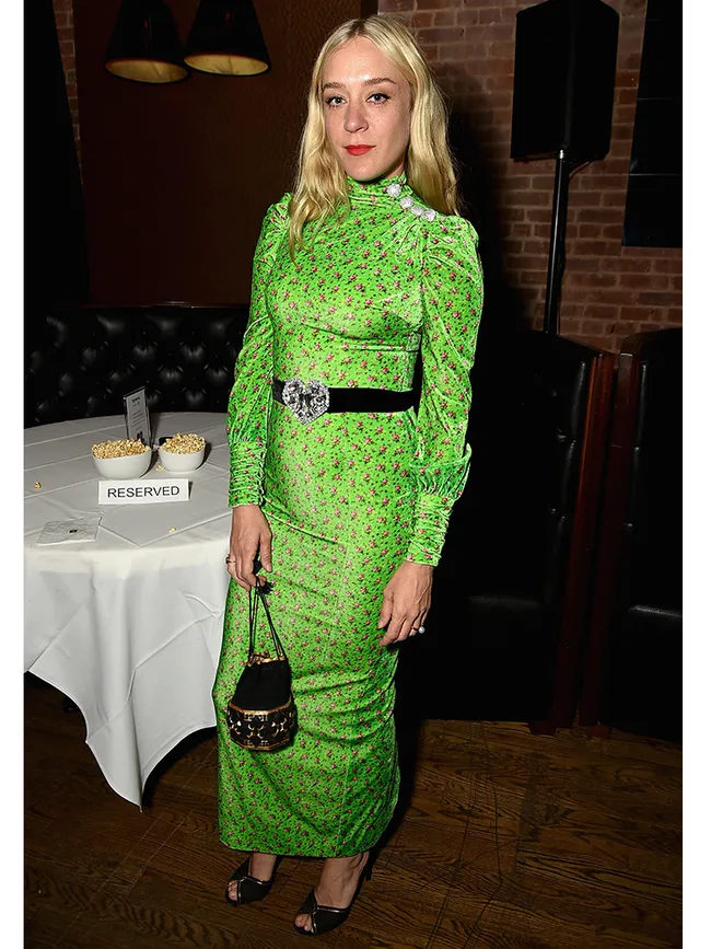 Blonde woman in a green floral dress with a black belt stands in a restaurant setting, holding a small handbag.