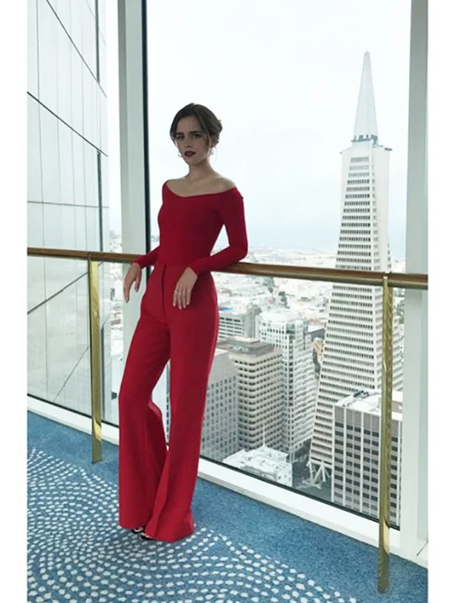 "Person in a red outfit standing in front of a cityscape with tall buildings visible through large windows."